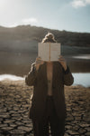 Woman in a tailored suit holding an open poetry book over her face on cracked desert earth