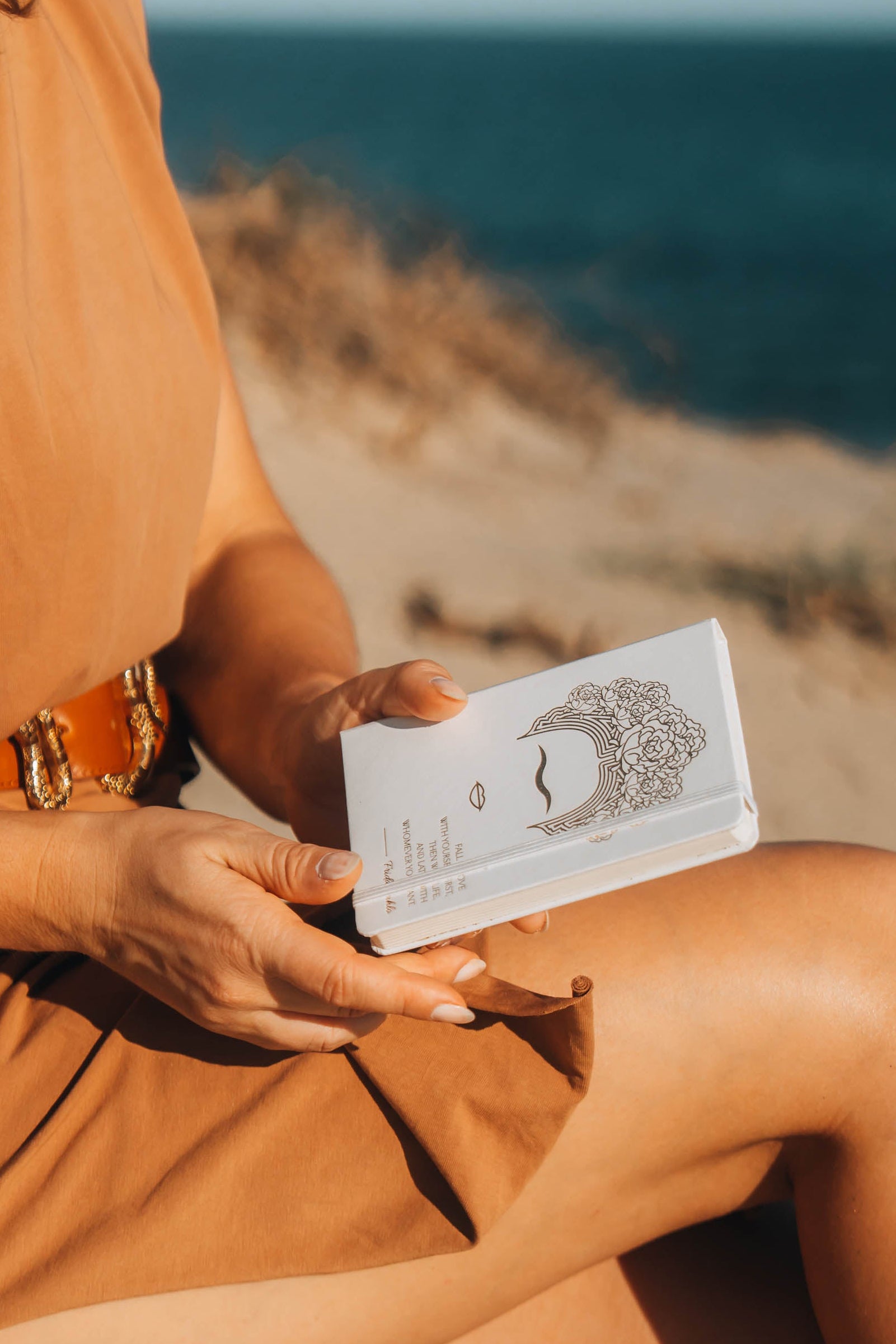 Woman in a brown dress sitting on a beach holding a book.