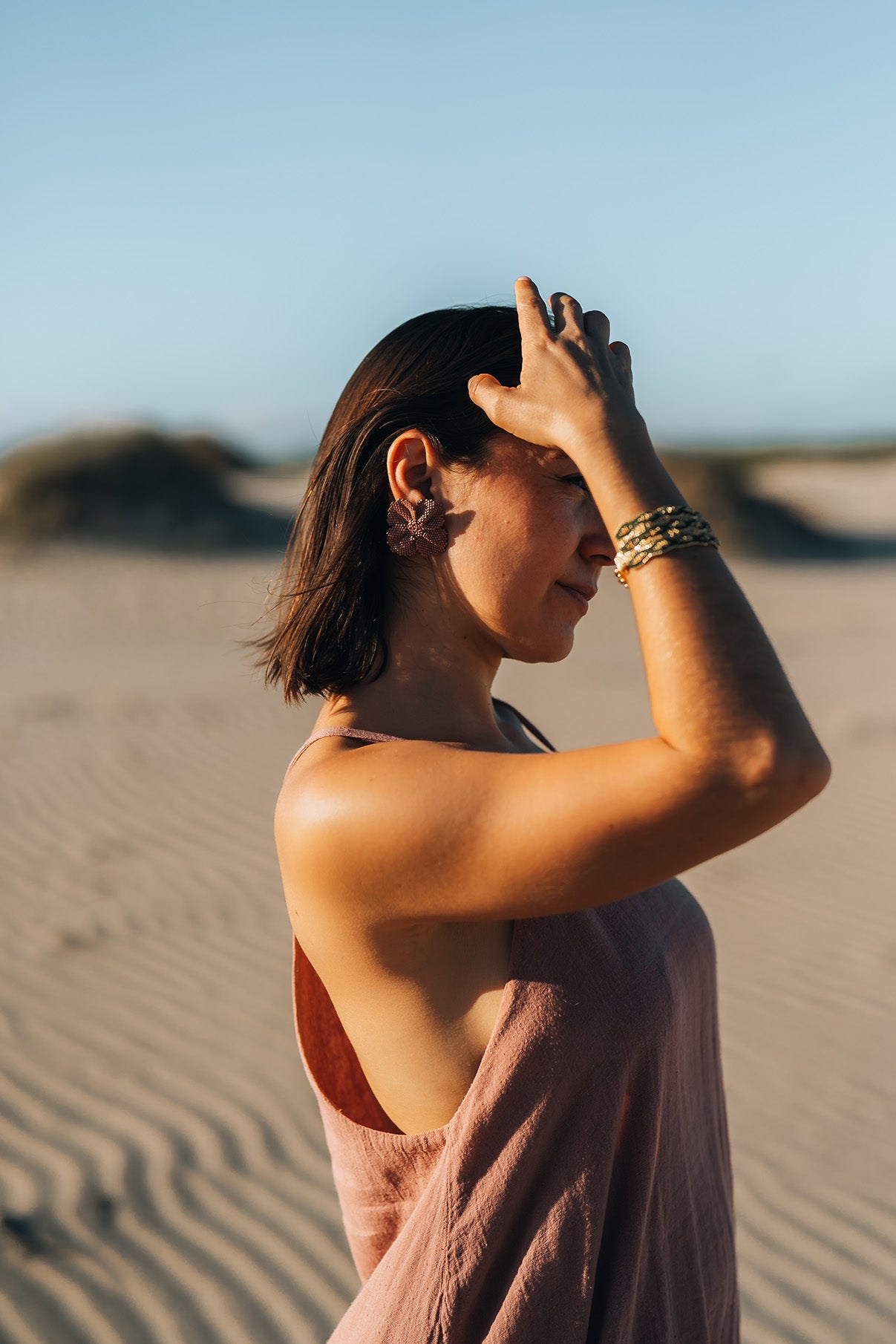 Woman standing on a sandy beach with a clear blue sky wearing flower earrings