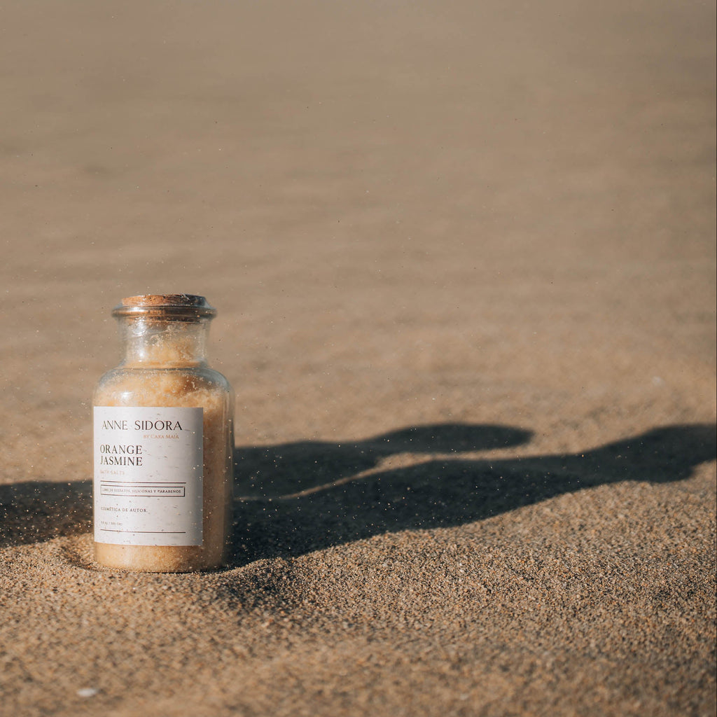 Two small bath salts bottles labeled 'Black Lavender' and 'Orange Spice' on a sandy surface.