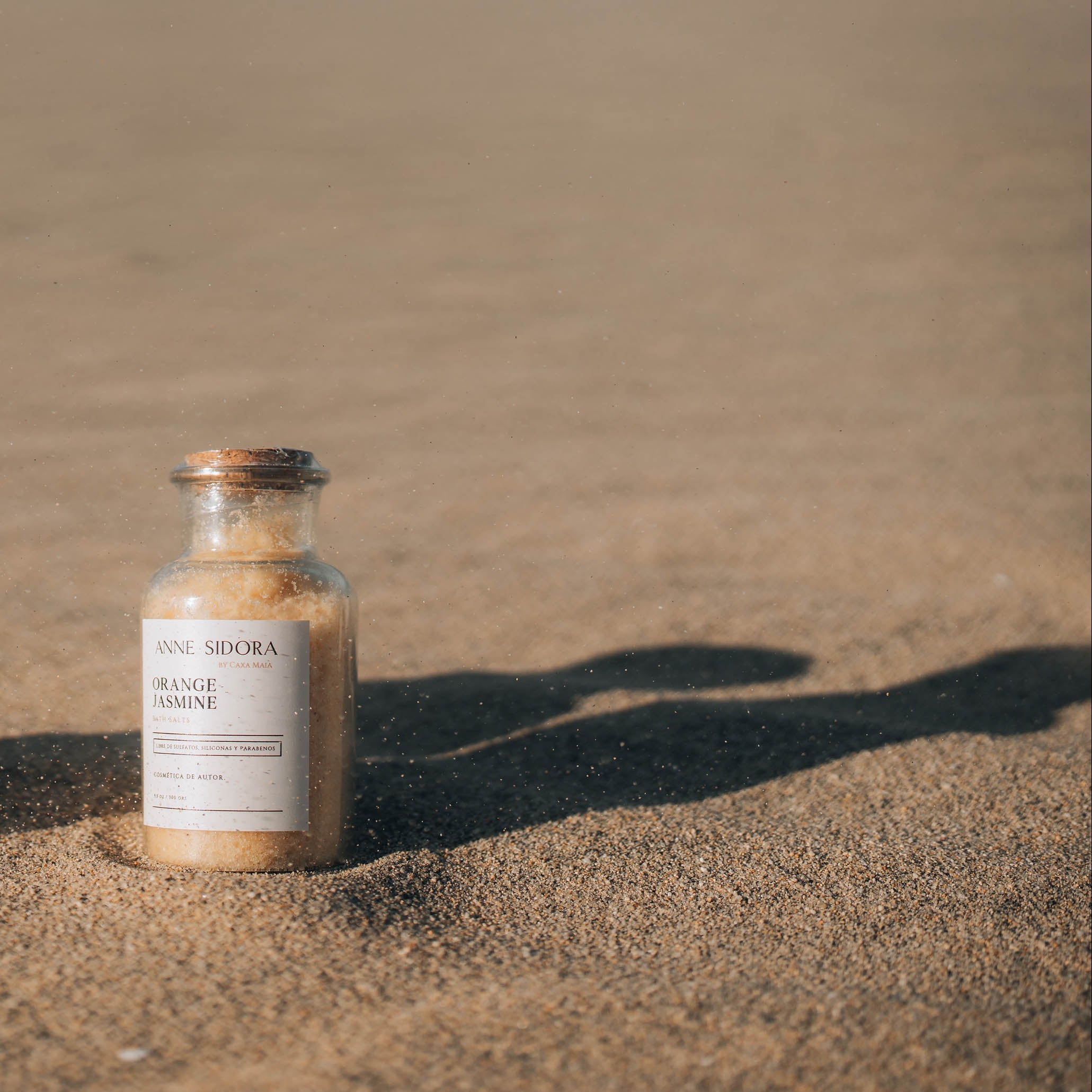 Two small bath salts bottles labeled 'Black Lavender' and 'Orange Spice' on a sandy surface.